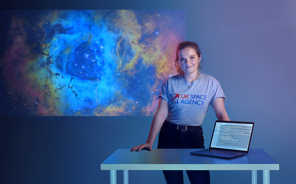 A young woman in UK Space Agency T-short stands next to a desk.
