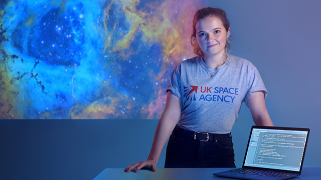 A young woman in a UK Space Agency t-short stands behind a desk.