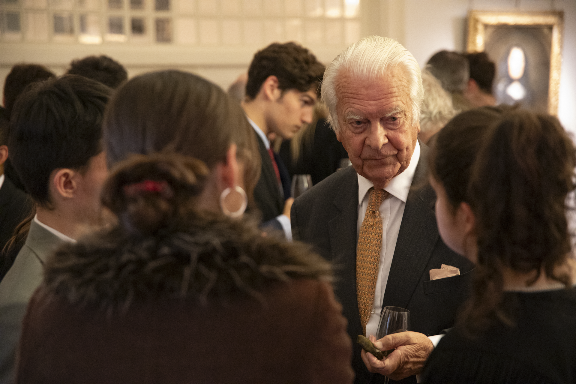 An older man (Lord Owen) speaks to a small group of students at a reception.